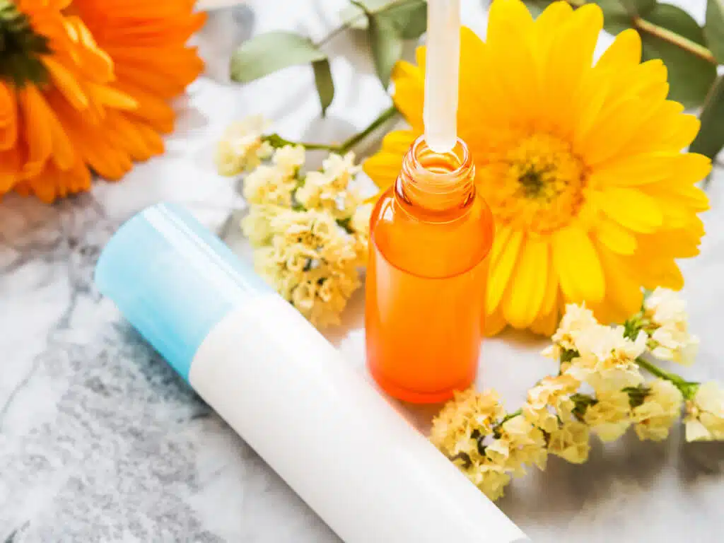 Face skin care products - white pump dispenser and anti aging serum in orange glass bottle on marble table with yellow flowers. Showing serum texture on dropper