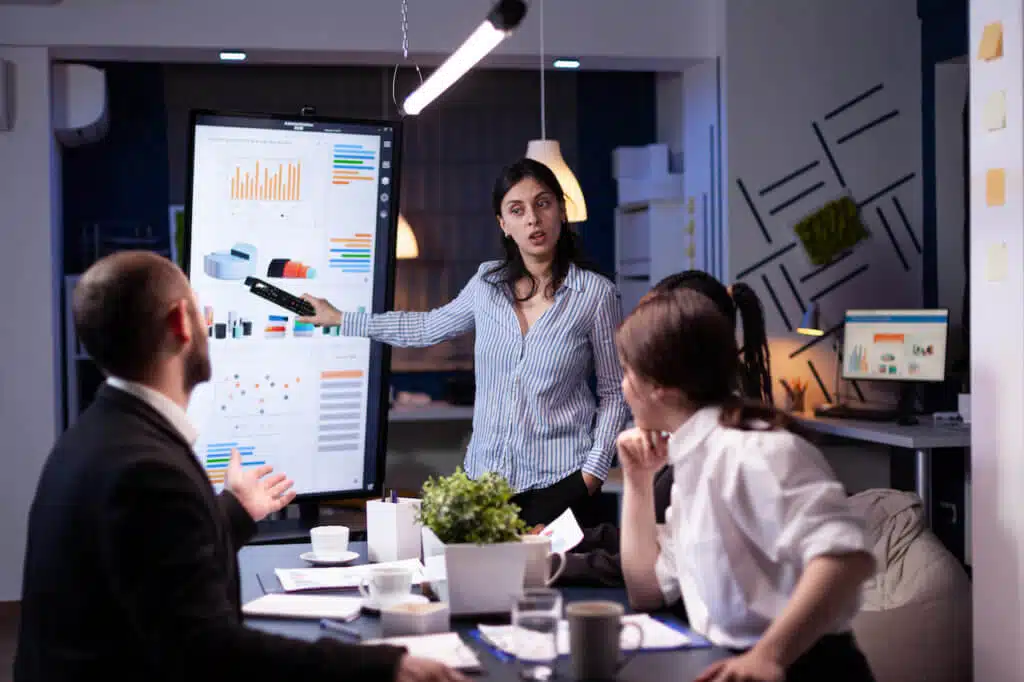 Workaholic focused businesswoman explaining management solution pointing strategy on monitor overworking in company business office meeting room. Multi-ethnic coworkers discussing ideas in evening. product lifecycle
