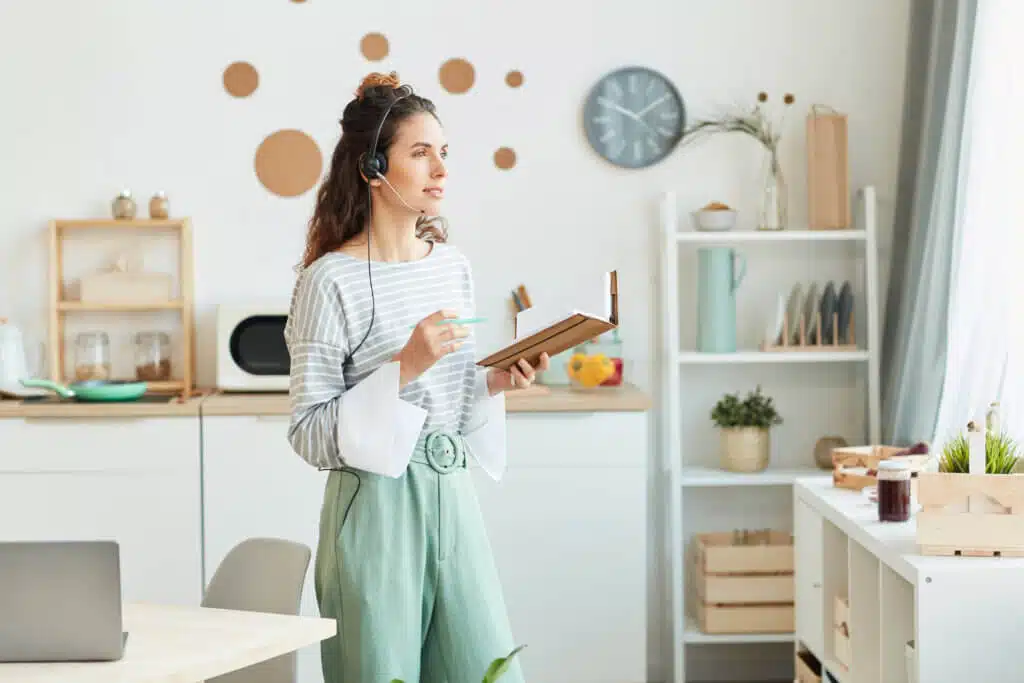 Caucasian woman standing in her kitchen at home holding notebook and pen working together with her colleagues during online meeting