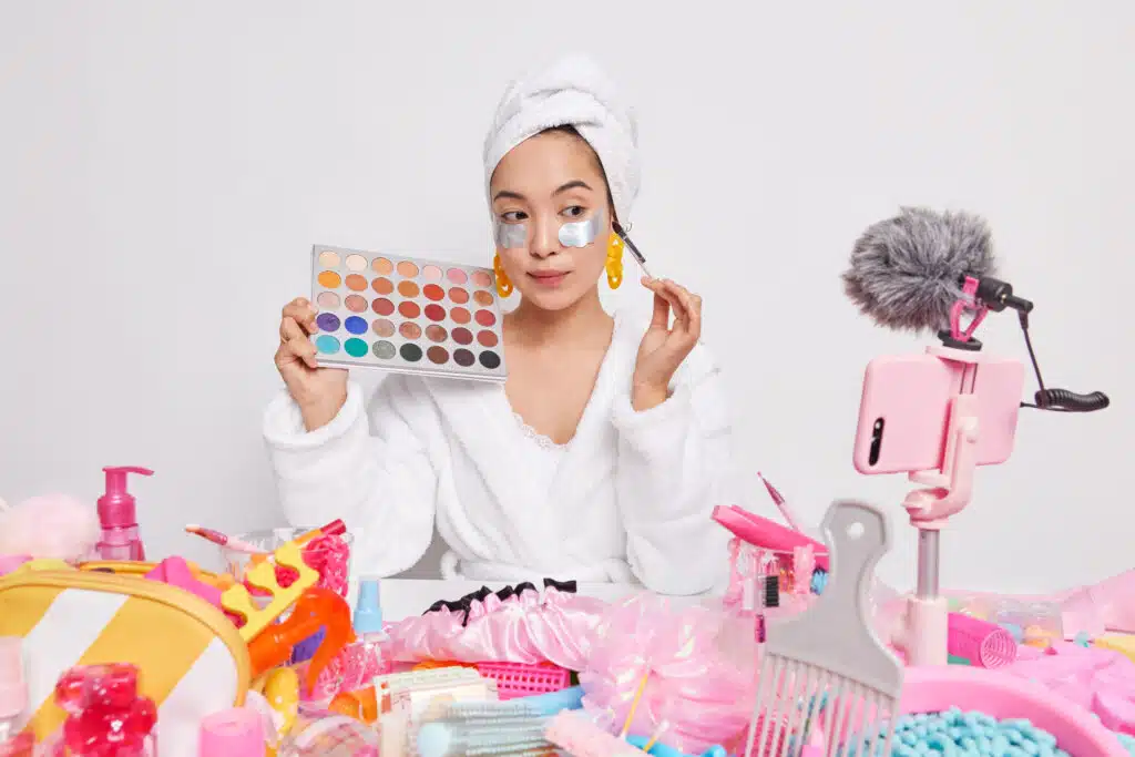Female beauty blogger tests cosmetics at home holds eyeshadow palette shares impressions with followers gives recommendations how to do makeup films process on smartphone webcam for viewers.