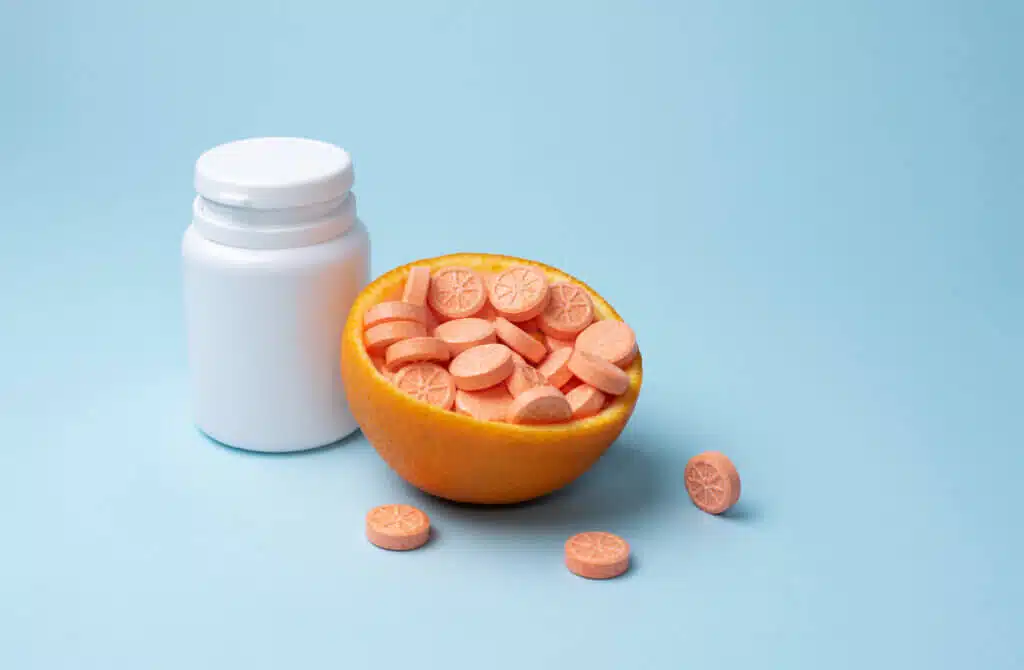 White bottle and orange tablets of vitamin C with half an orange on a blue background, mockup. Immune booster concept.