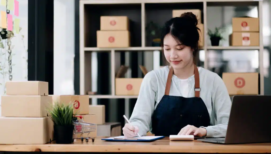 A portrait of a young Asian woman, e-commerce employee sitting in the office full of packages in the background write note of orders and a calculator, for DTC business, e-commerce and delivery business of D2C trends
