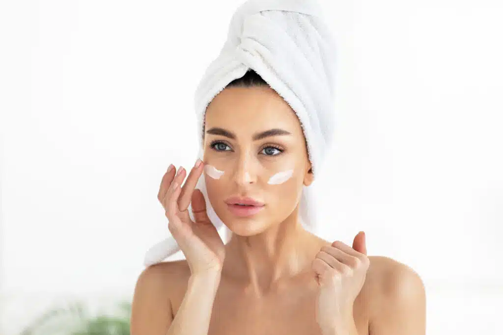 Clean skin cosmetics, dermatology care, anti-age mask and natural beauty. Young european cute woman in towel applies moisturizer cream on face at home, on white bathroom background, copy space