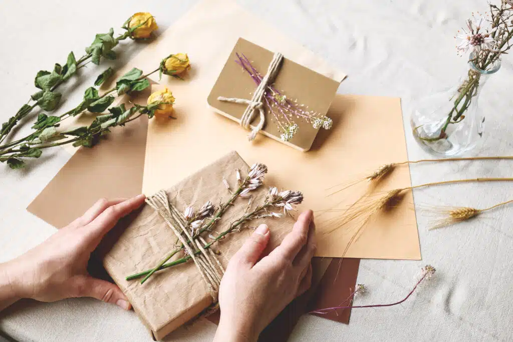 Woman makes zero waste, plastic free, trendy hand made gift package with craft recycled paper and dried flowers on the table with linen tablecloth. Natural aestetic.