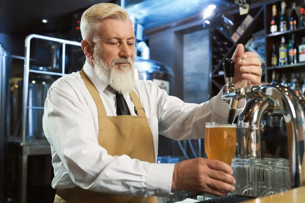 Handsome, bearded, elderly barman standing at bar counter. Bartender in white shirt, tie, brown apron pouring in cold glass lager beer.
