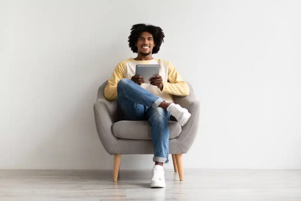 Cheerful Young Black Man Relaxing In Chair With Digital Tablet, Smiling Millennial African American Guy Using Modern Gadget While Resting At Home, Browsing Internet Or Shopping Online, Copy Space