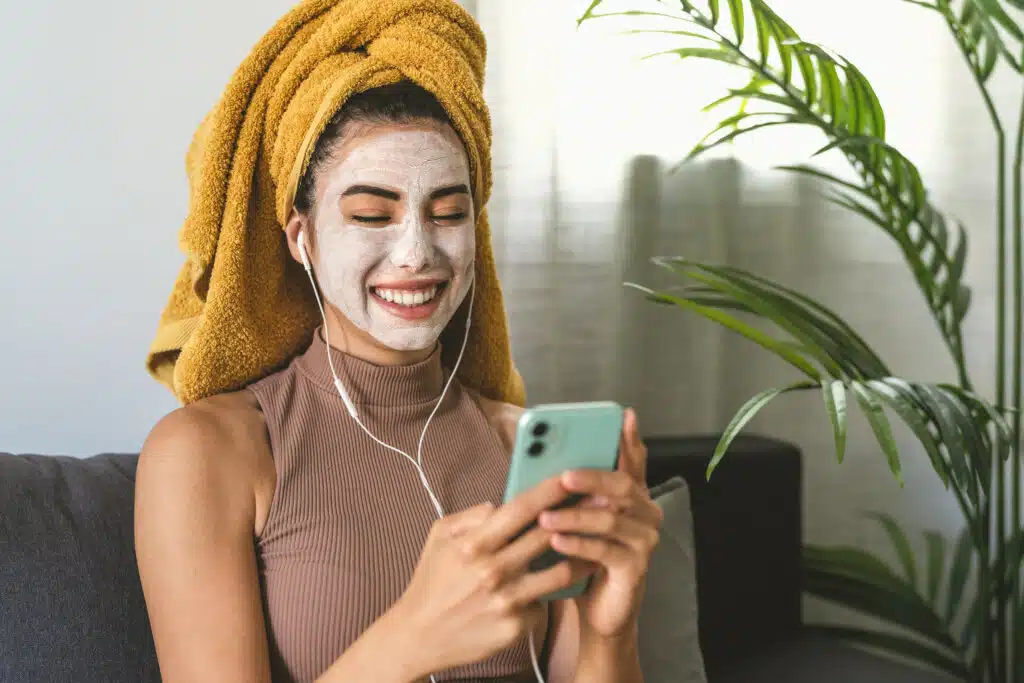 Young woman using mobile smartphone while having skin care spa day at home