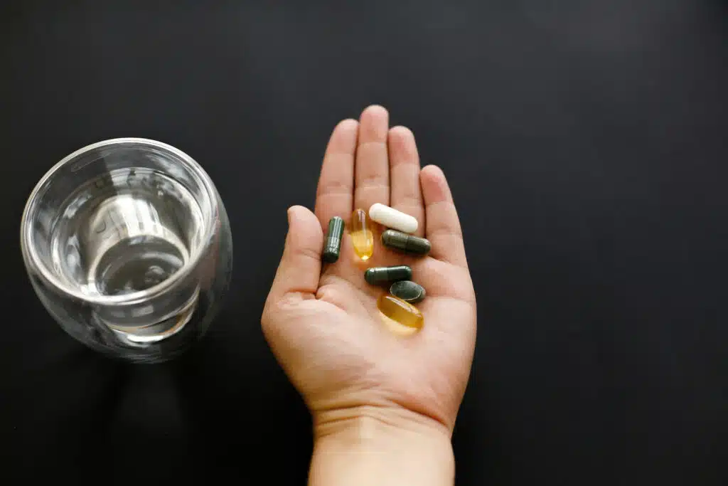 Hand holding omega 3, spirulina, chlorophyll,magnesium  capsules above glass of water. Morning dose of pills. Dietary supplements. Health support. Biologically active additives
