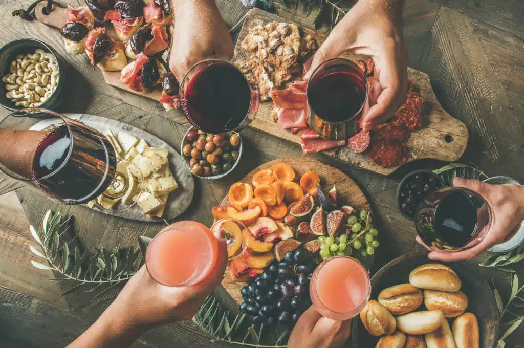 Flat-lay of friends eating and drinking together. Top view of people having party, gathering, dinner together sitting at wooden rustic table set with wine snacks and fingerfoods. Hands with glasses