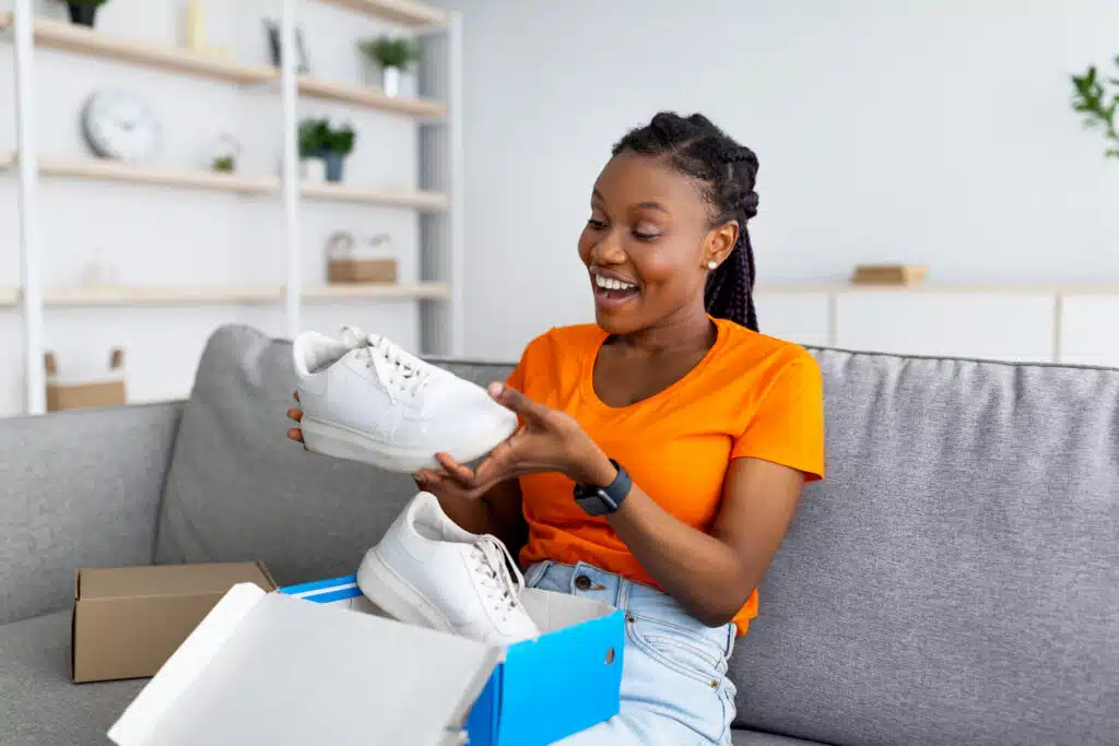 Satisfied black female client opening box from online shop and looking at delivered item, happy with received sneaker shoes, enjoying footwear after unboxing package at home