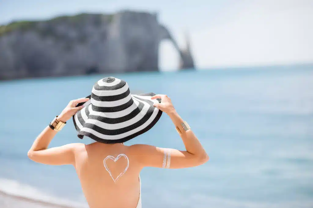 Back view on a beautiful woman in striped hat with sunscreen heart shape on her shoulder having a sunbath on the famous french beach