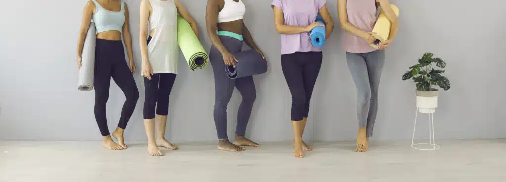 Group of beautiful fit women holding workout mats standing by wall in sports gym or yoga studio. Cropped low section shot of slim female legs in leggings and yoga pants. Fitness fashion concept banner