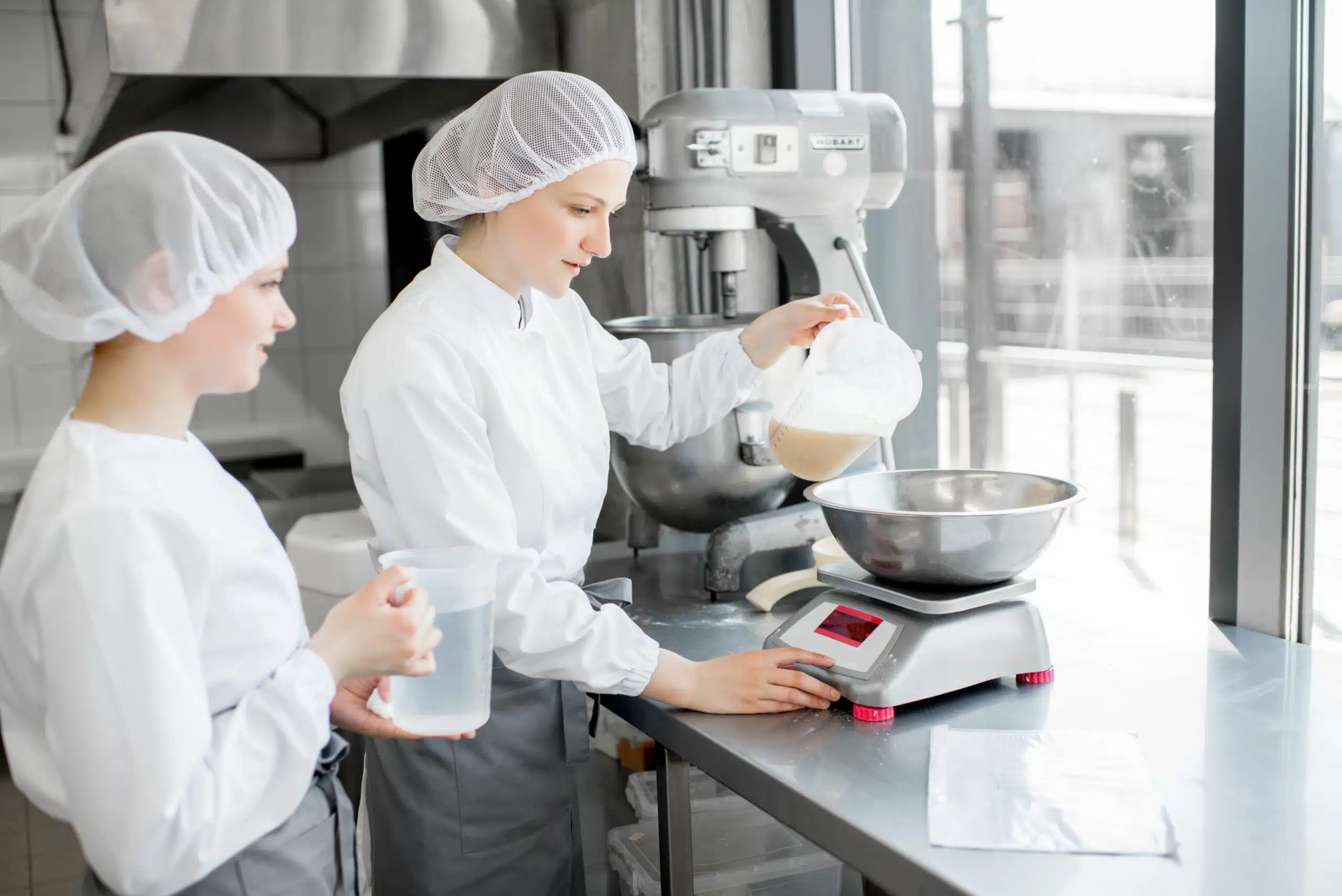 Two female confectioners in uniform weighing ingredients for pastry working at the bakery manufacturing
