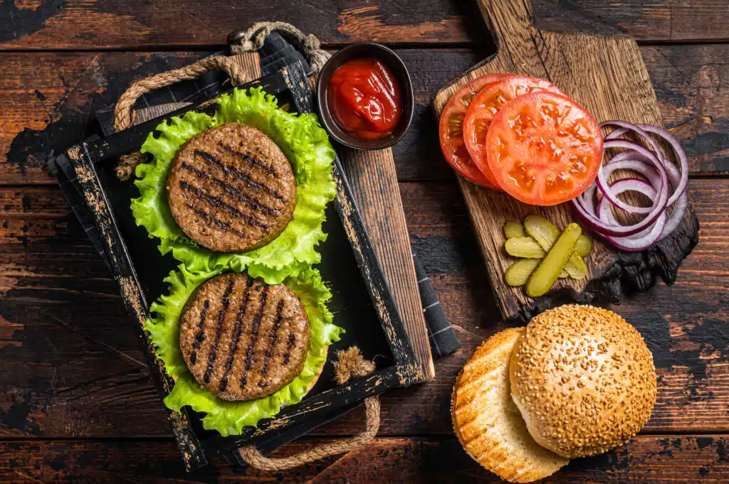 Cooking Plant based meatless burgers with vegetarian meat free roasted cutlets, patties, tomato and onion in a wooden serving tray. Wooden background. Top view.