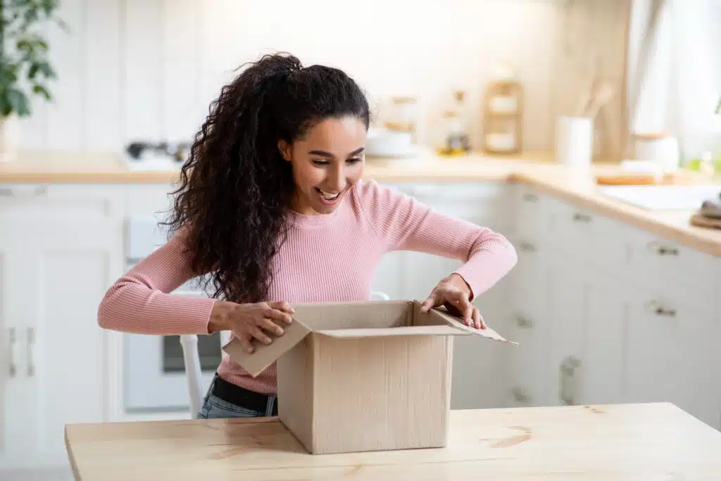 Excited happy young woman unboxing cardboard parcel In kitchen, millennial female got box delivery after successful online shopping in internet, cheerful lady received her order, copy space