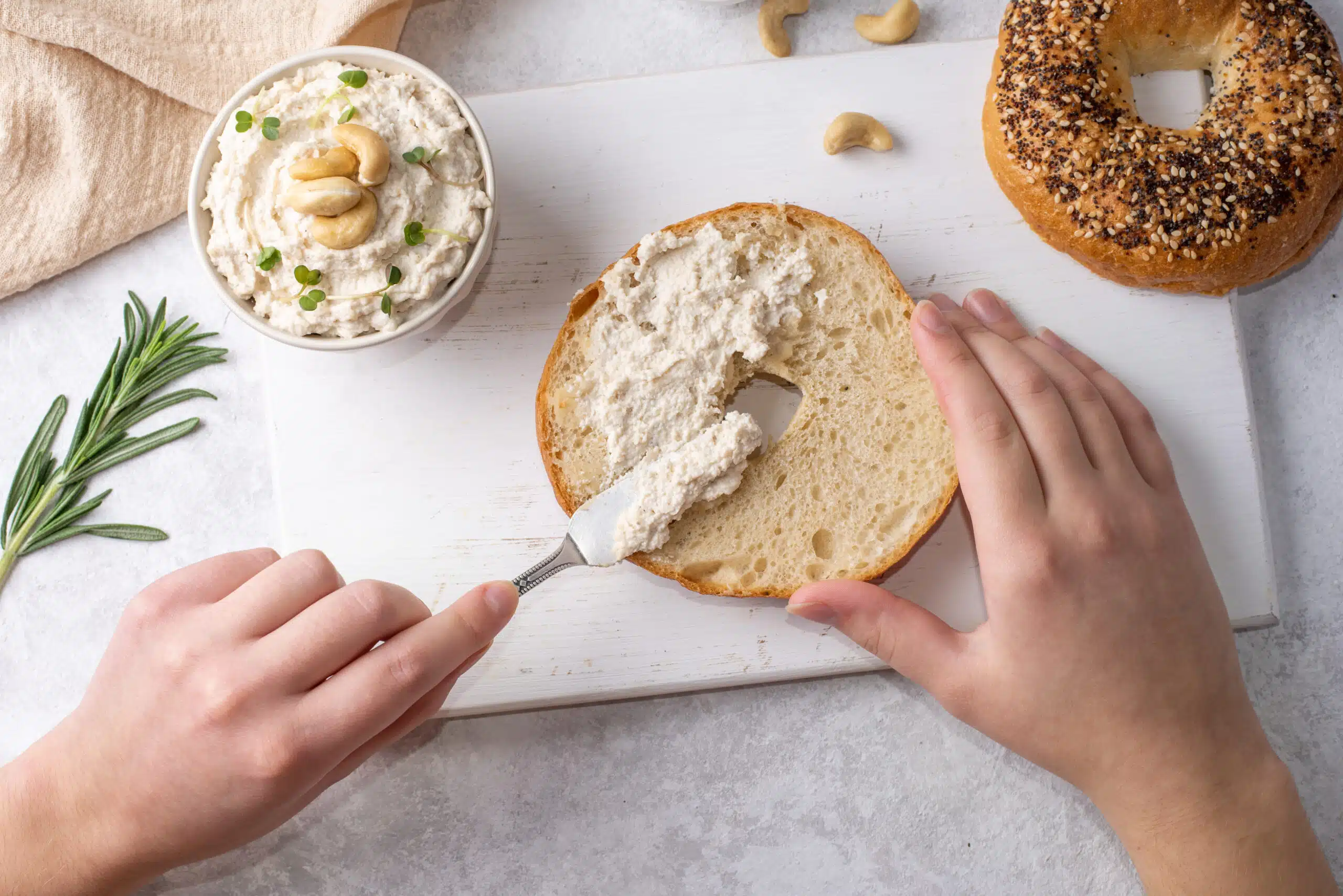Female hands spreading cashew cheese on a bagel on a white table, fermented nuts as an alternative to cream cheese.