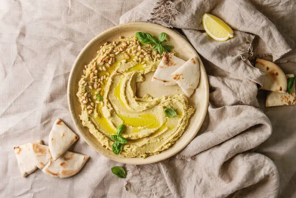 Homemade traditional spread hummus with pine nuts, olive oil, basil served on ceramic plate with pita bread over linen cloth background. Flat lay, space. Mediterranean snack.