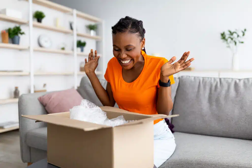 Overjoyed Afro woman unboxing carton parcel, emotional about successful shopping, satisfied with great purchase at home. Online store, reliable delivery service concept