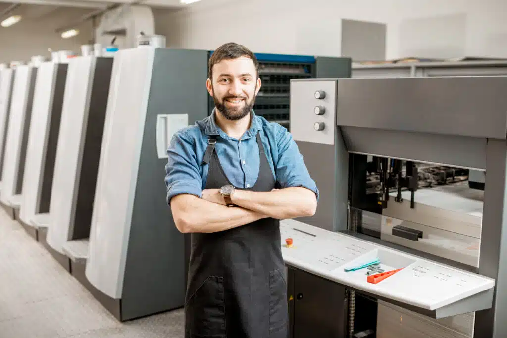 Portrait of a handsome typographer standing at the printing manufacturing with offset machine on the backgorund