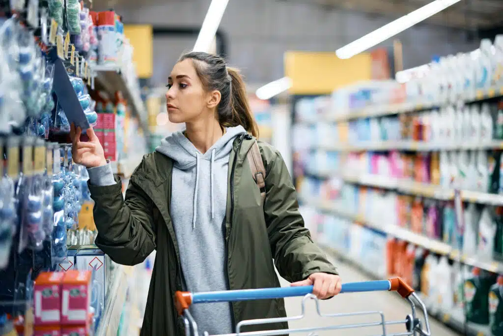 Young woman shopping supplies in supermarket and looking at product prices.
