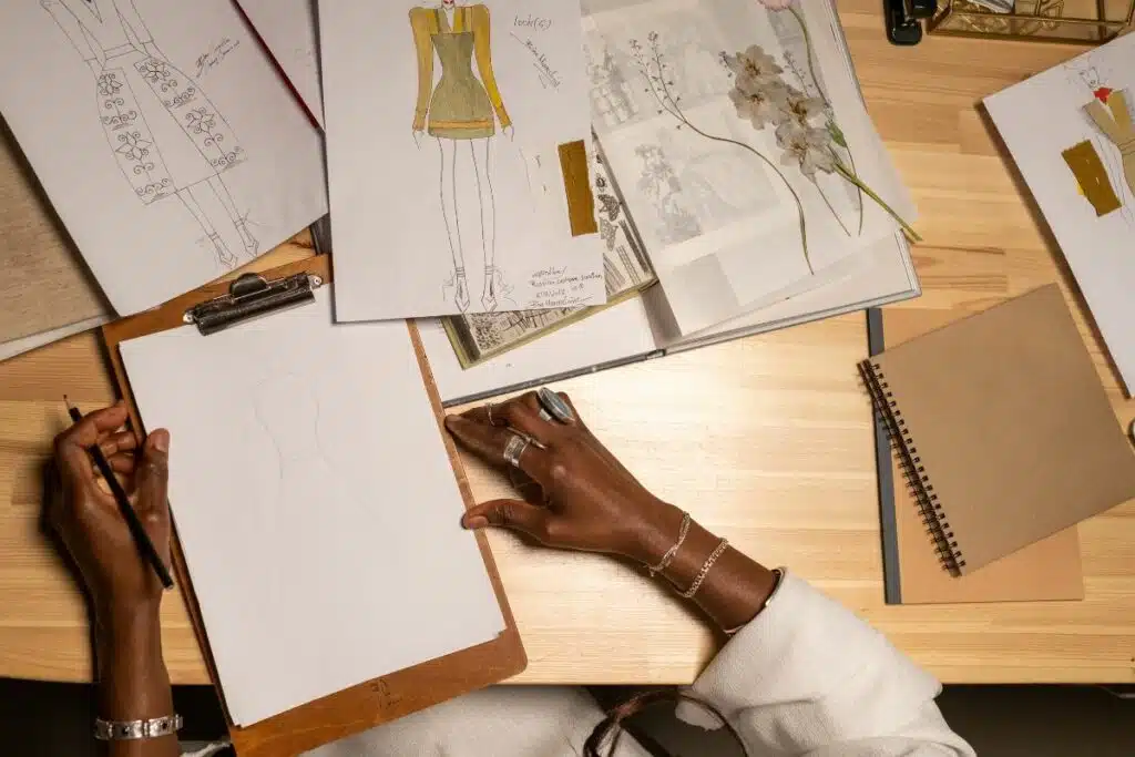 A focused woman, with fashion sketches and fabric swatches spread before her, meticulously planning a clothing line, showcasing the creative process behind a fashion brand's collection.